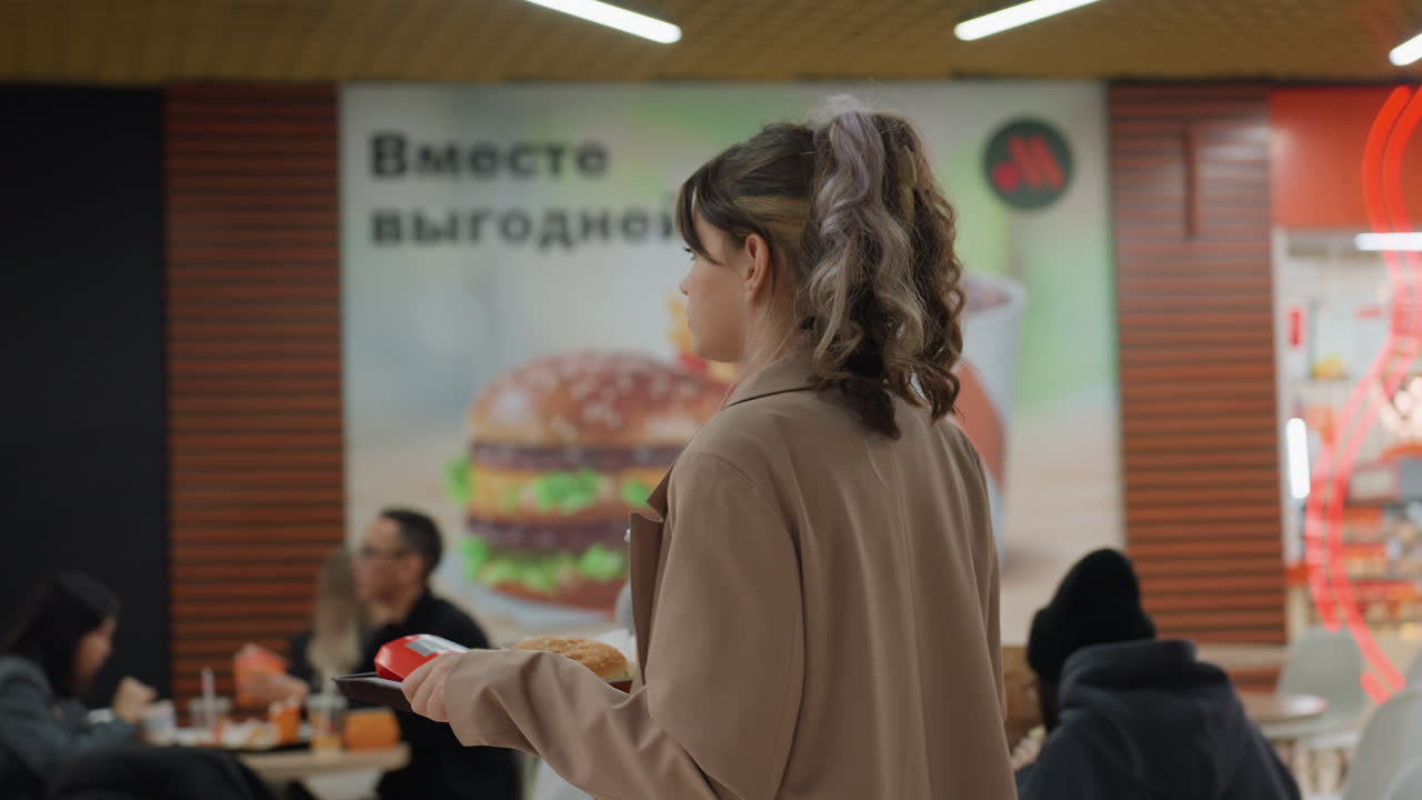 쇼핑하는 젊은 여성, 쇼핑몰에 있는 여성, 식당 메뉴를 살펴보는 젊은 여성, 붐비는 푸드 코트 통로를 다니는 백인 여성, 붐비는 쇼핑몰 식사 공간을 탐색하며 쟁반을 들고 있는 젊은 금발 여성