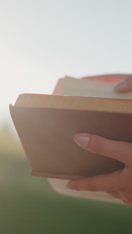 la luz del sol brilla de la vegetación borrosa en el fondo, reflejándose en un libro sostenido por una mujer sentada al aire libre, ella revisa suavemente las páginas mientras lleva gafas en su camiseta negra