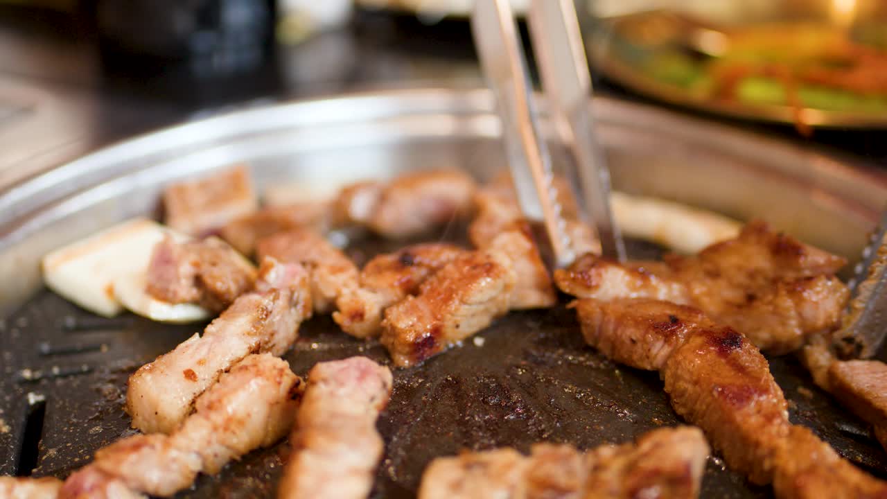 Grilled pork belly pieces turned with metal tongs on a round tabletop Korean barbecue grill