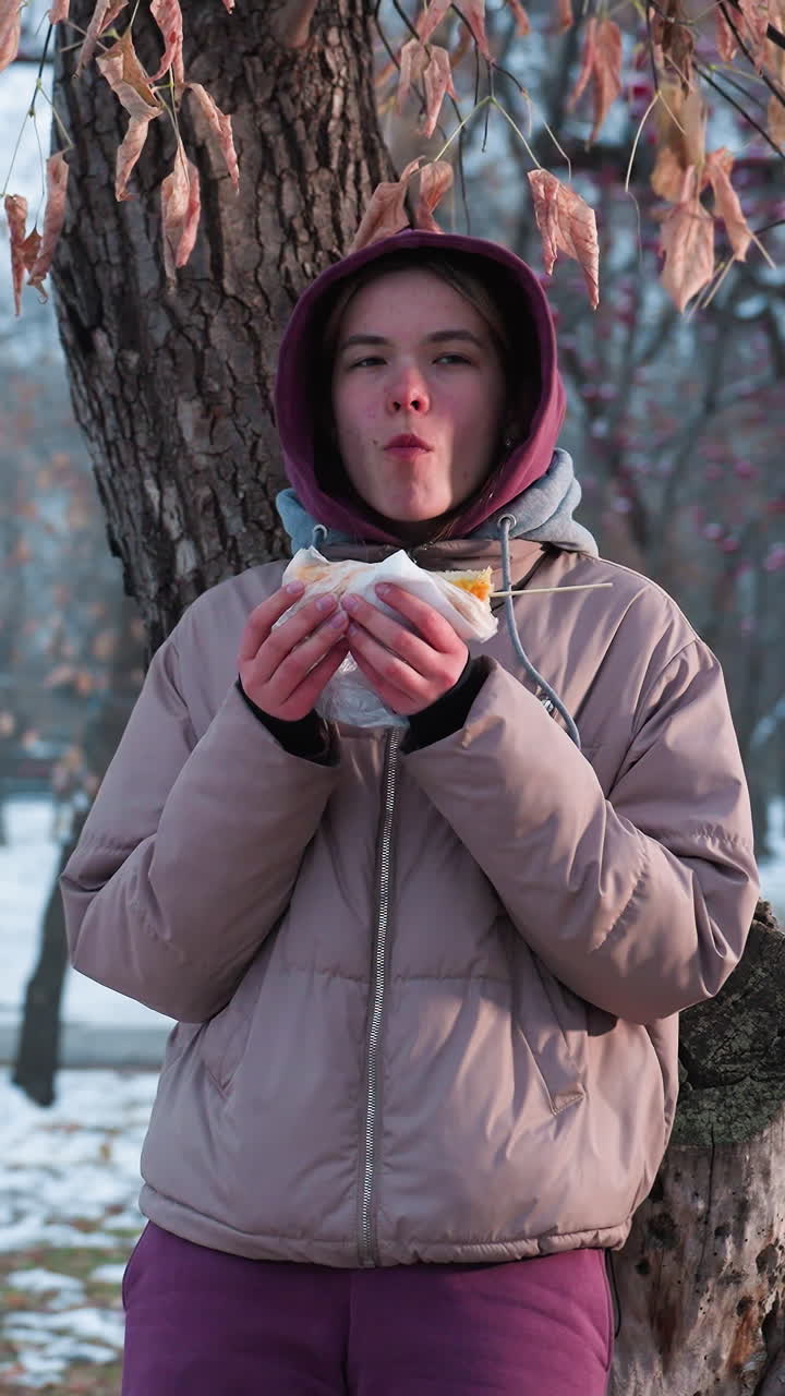 mujer joven en traje de invierno descansando en un árbol comiendo un bocadillo en un parque al aire libre durante el invierno, reflejo de luz desde el fondo, disfrutando del momento de la comida, acogedor descanso de invierno en la naturaleza