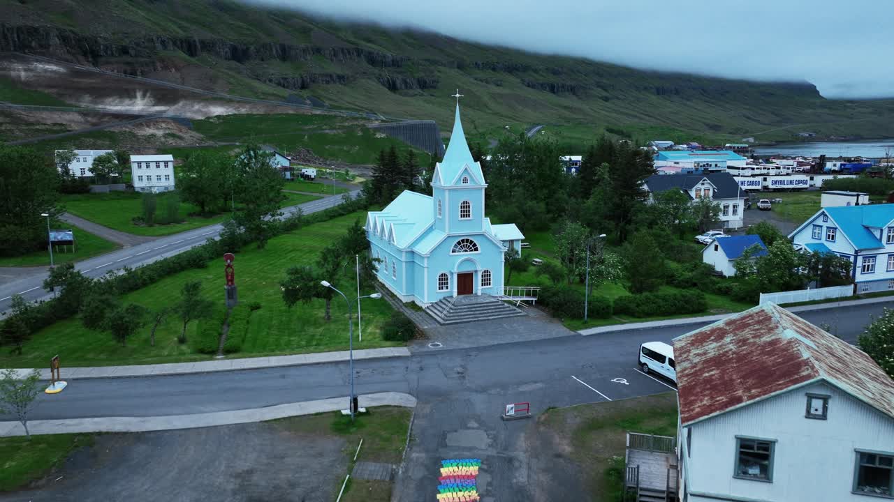 Aerial approaching shot of blue church in seydisfjordur town during cloudy day. Parking cars and green mountain coastline in background