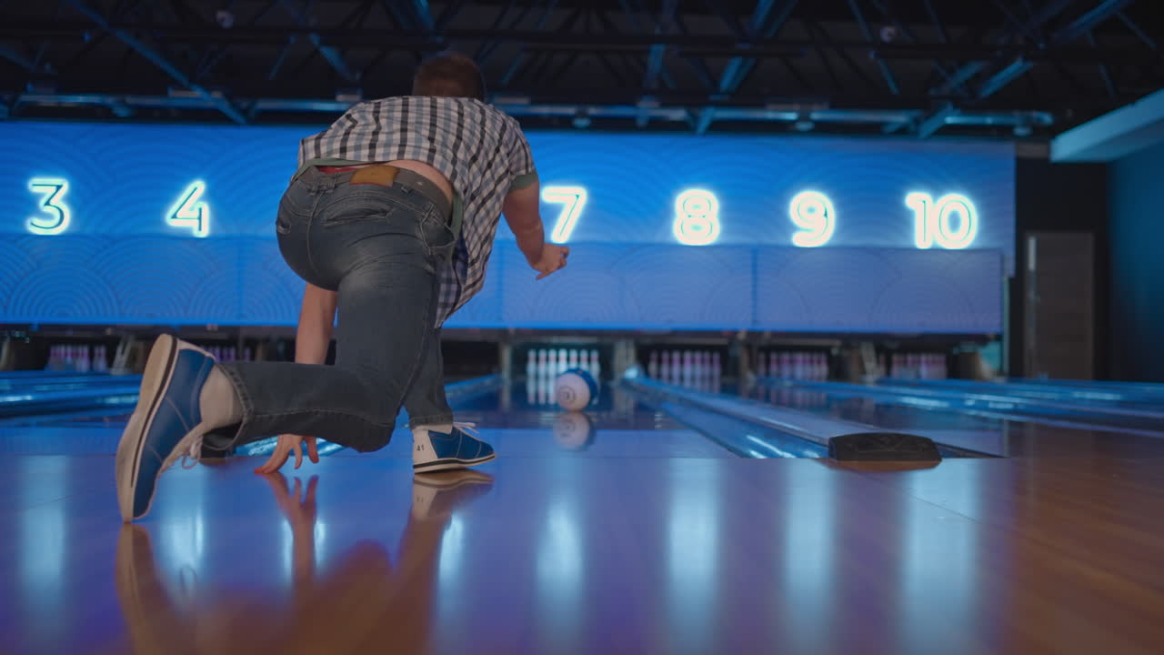 a câmera segue um homem caucasiano jogando uma bola de bowling na pista de jogo e saltando alegres nos pinos caídos. um homem jogando bowling em câmera lenta