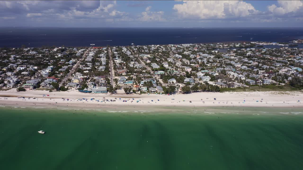 Rows of coastal homes stretch beside a broad white sandy beach where calm turquoise waves meet the shore, capturing the relaxed energy of seaside living under bright daylight