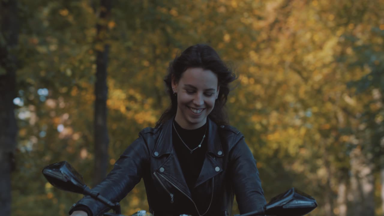 Pretty smiling European young woman driving a motorbike wearing leather jacket in forest with vibrant, colorful golden autumn leaves on sunny day-5
