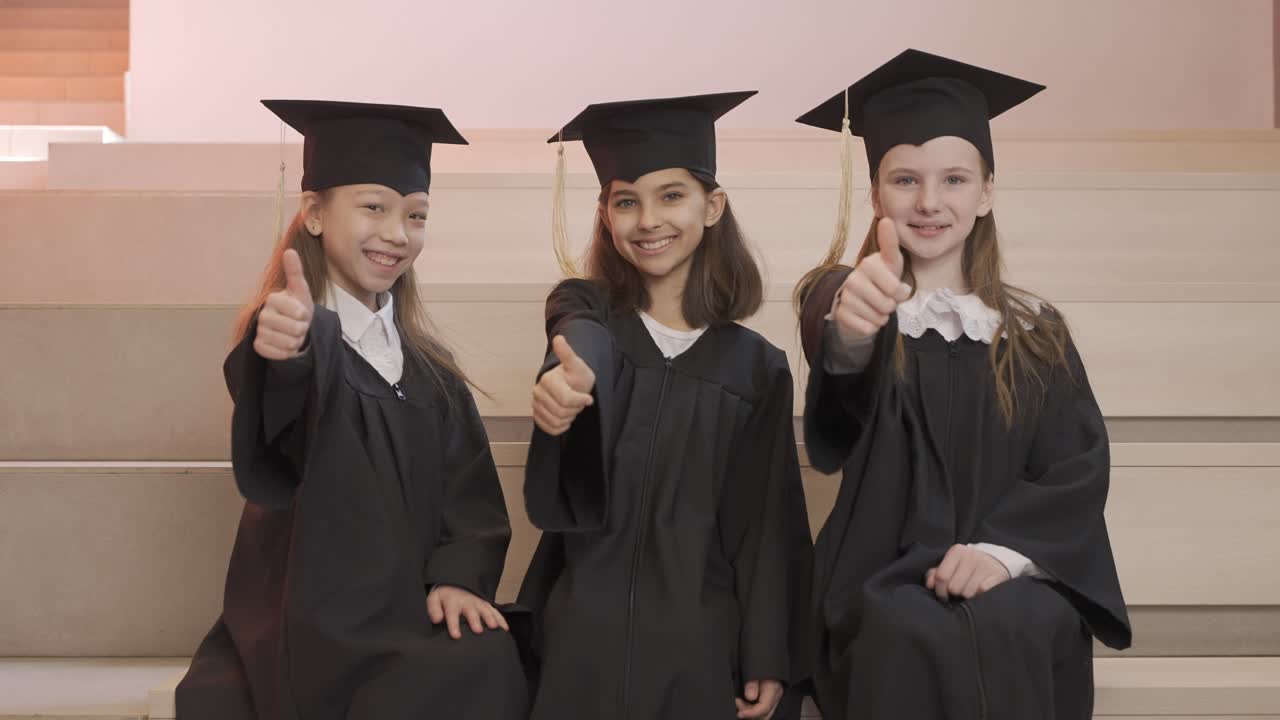 увеличение изображения счастливых воспитанников детского сада в шапочках и мантиях, смотрящих в камеру и делающих знак "большой палец вверх" во время церемонии вручения дипломов в детском саду