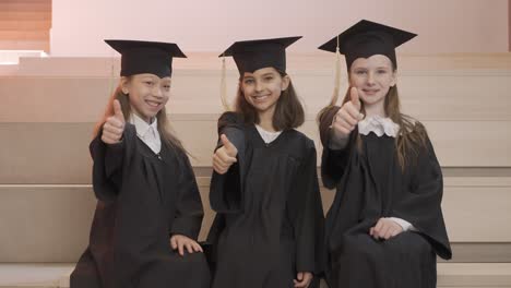acercar a los felices estudiantes de jardín de infantes con toga y birrete mirando la cámara y haciendo el signo del pulgar hacia arriba durante la ceremonia de graduación del preescolar