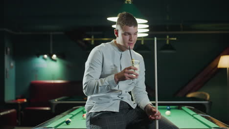 billiard player in white shirt and grey trousers sits on green pool table holding cue stick and sipping lemon drink. pool hall setting with stylish overhead lighting and relaxed atmosphere