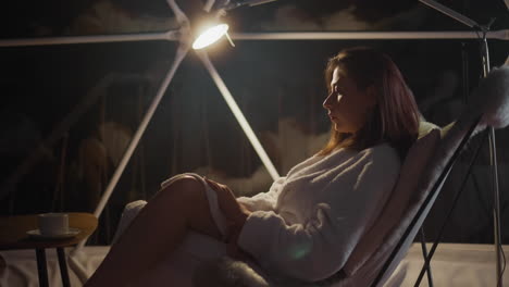 gloomy lady covers legs with terry robe sitting in armchair by light of lamp near panoramic window at night. lack of support leading to suffering from loneliness slow motion