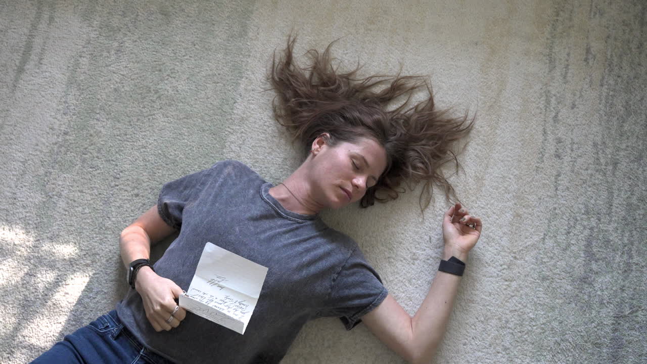 A gentle scene of a woman lying on the ground, holding a love letter on her stomach, as the camera slowly zooms out and rotates around her.