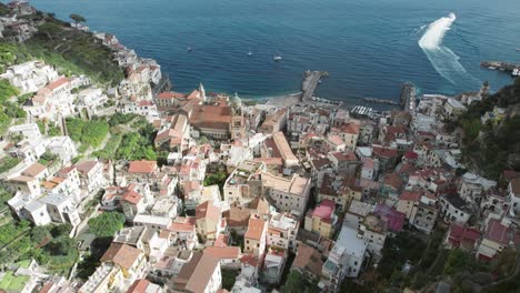 An-aerial-shot-of-a-boat-departing-the-port-of-Amalfi-and-showing-the-rustic-slate-roofs-of-historic-Amalfi-town,-one-of-the-most-popular-tourist-destinations-on-the-famous-Amalfi-Coast-in-Italy