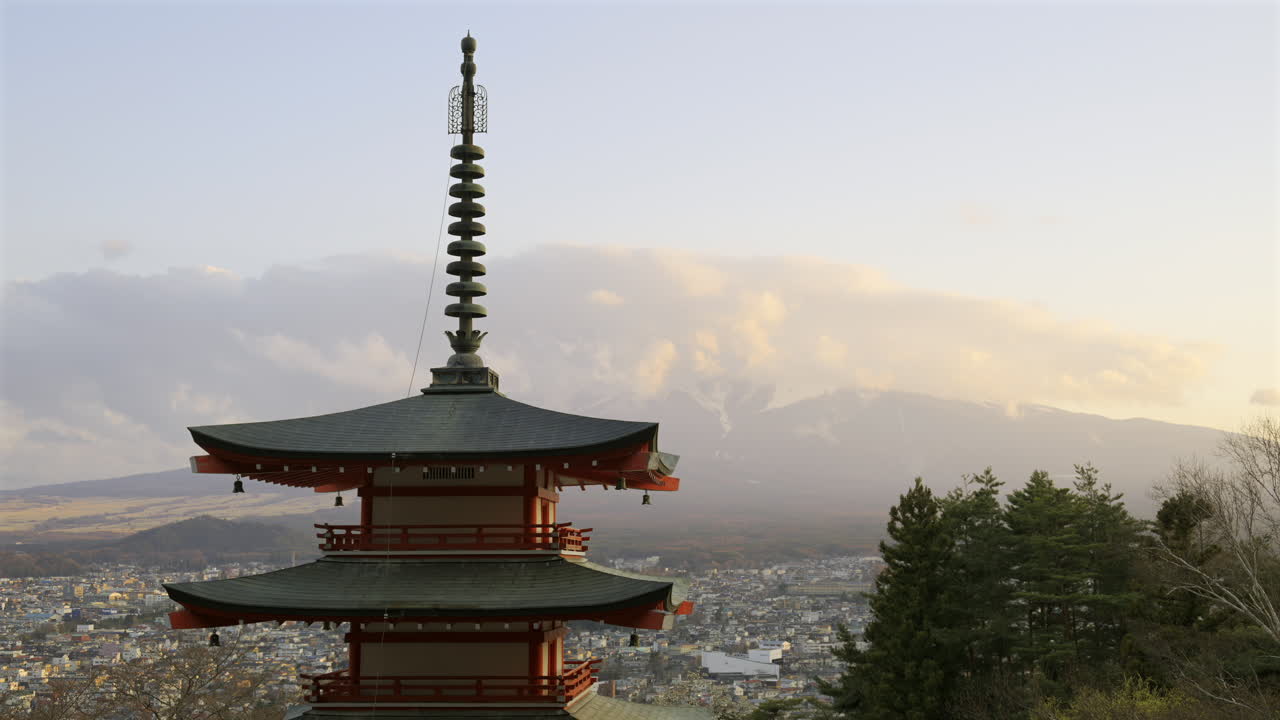 Chureito Pagoda stands tall, overlooking a sprawling Fujikawaguchiko city as the sun sets behind majestic Fuji mountain. The colorful architecture contrasts beautifully with the evening sky