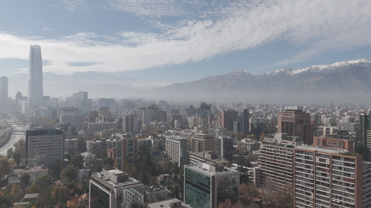 Forwards drone shot of Santiago de Chile in South America watching over the skyscrapers and big buildings with snowy mountains in the background on bright but cloudy day LOG