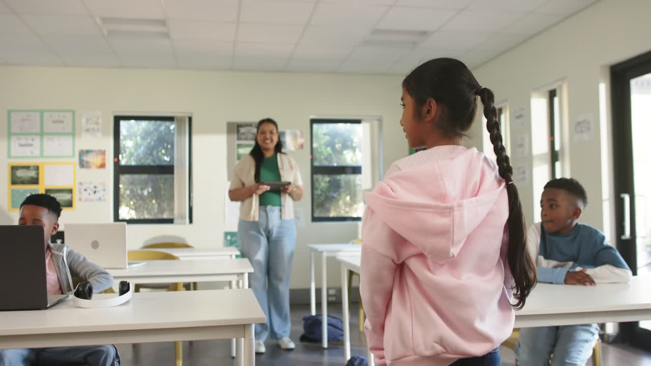 Teenage girl in pink hoodie walking to front in class after teacher with tablet smiling presenting
