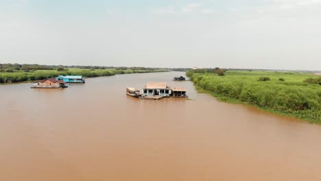 drone flying towards four floating buildings with a boat and inbetween green shoreline