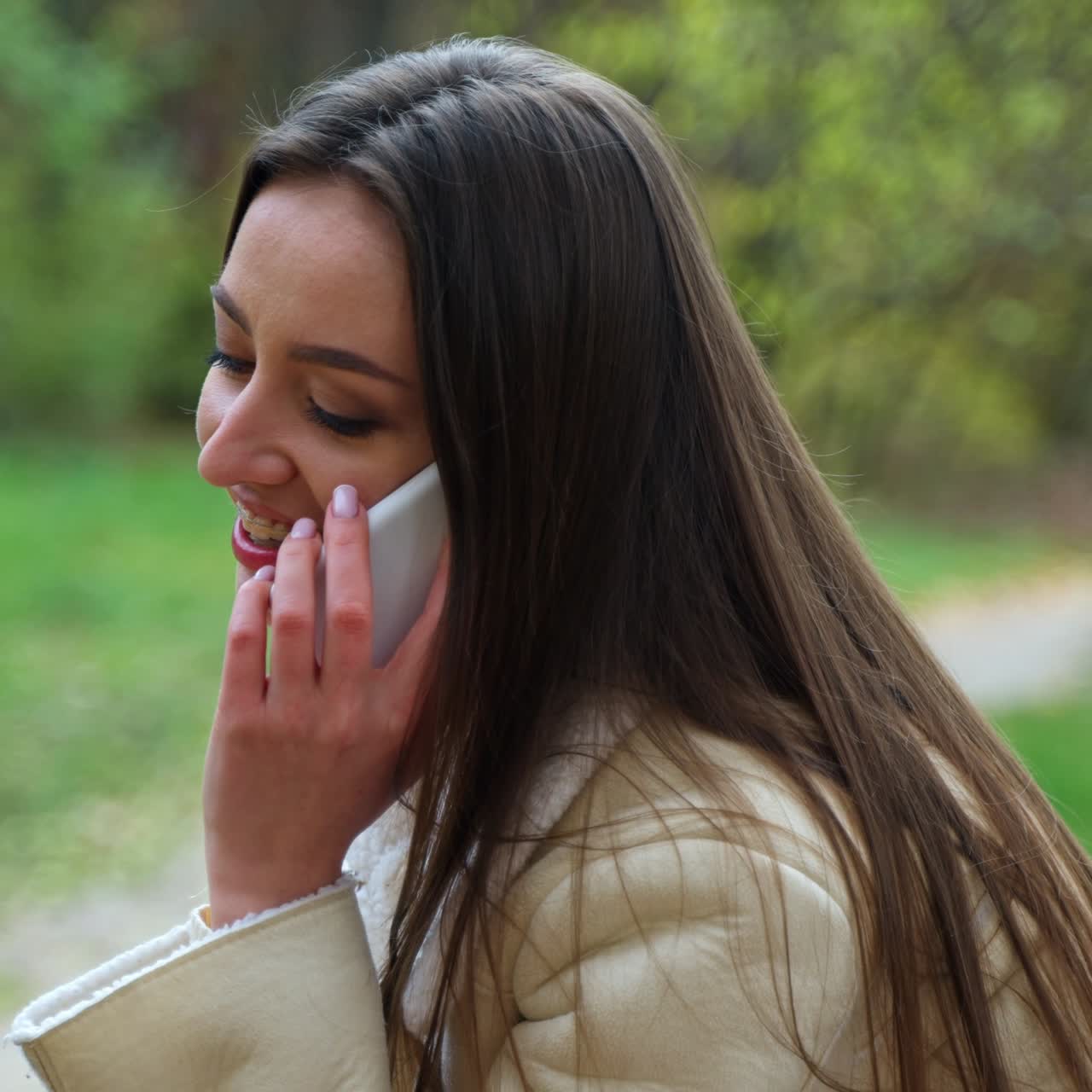 Young woman having phone conversation sitting in the nature. Lady with straight long hair laughing happily during her talk. Blurred autumn backdrop