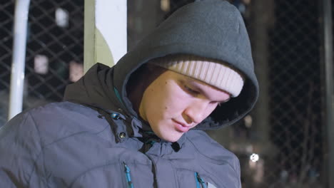 close-up of young man in hoodie and beanie reading a red cover book outdoors at night, illuminated by urban city lights with mesh fence in the background