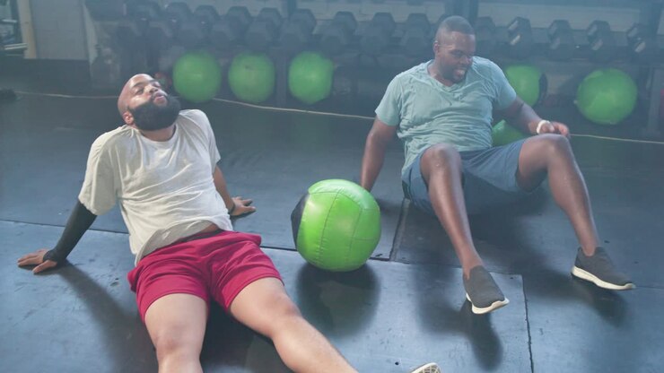 Two African American men holding green medicine ball, rotating and passing for core workout at gym