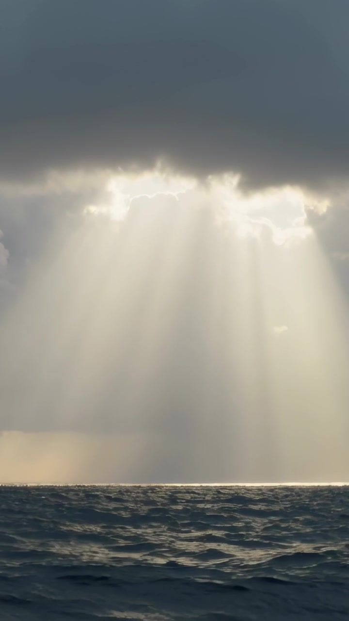Vídeo vertical: Colunas de luz solar perfurando uma fenda nas nuvens sobre o mar, formando uma faixa brilhante no horizonte.