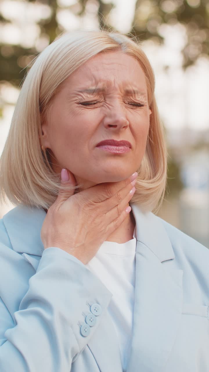 Sick mature businesswoman in formal suit suffering from sore throat while standing on city street