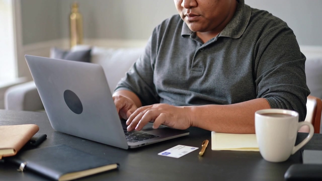 Reaching for card, middle-aged Hispanic man typing on laptop at home, entering payment details