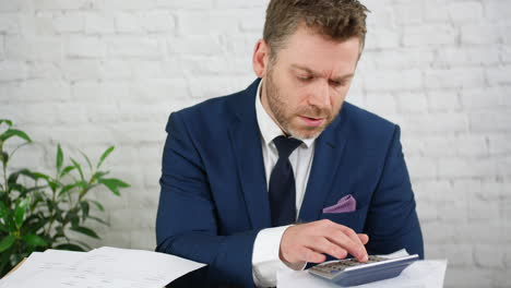 a stressed businessman using a calculator calculating debts and taxes with piles of financial accounts paperwork in an office