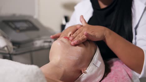 young woman is lying on the couch during cosmetic face procedure. professional cosmetologist is making face massage in spa salon