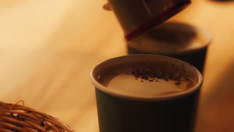 Close-up-of-cocoa-powder-being-sprinkled-on-a-coffee,-in-a-paper-cup,-inside-a-coffee-shop,-slow-motion