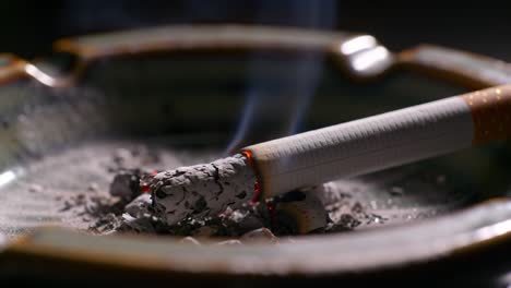 close-up of a smoldering cigarette in an ashtray, capturing the glow of embers and the surrounding ashes in a dimly lit environment