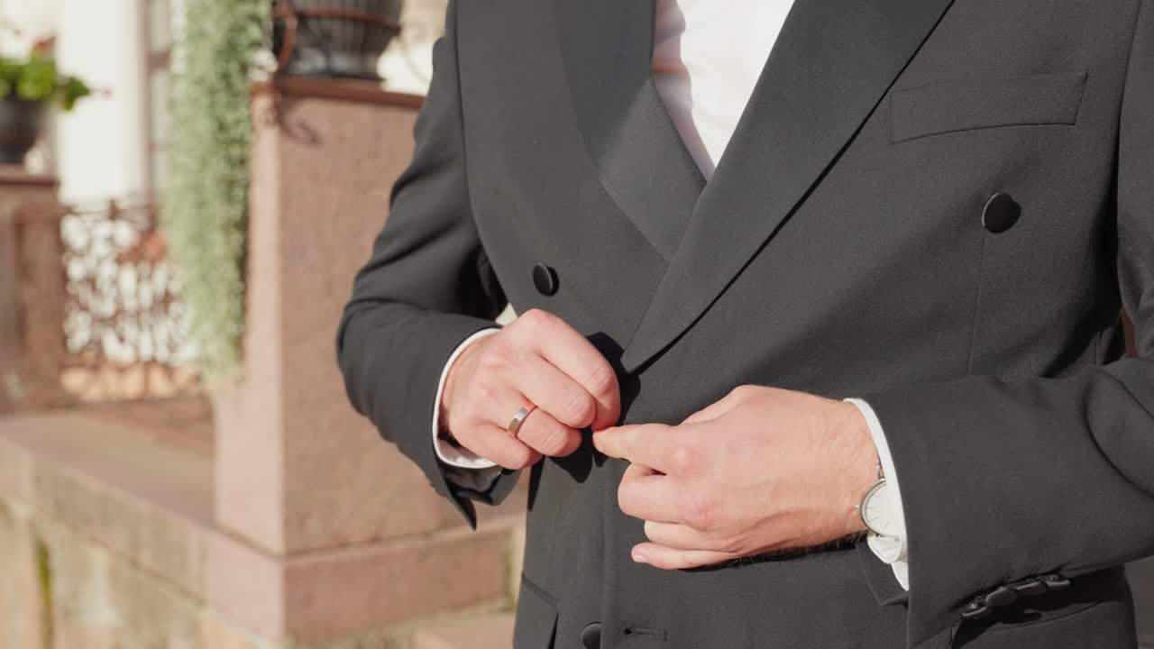 Un uomo che si abbottona meticolosamente la giacca del suo abito nero formale in un'elegante ambientazione esterna. La luce solare brillante evidenzia il taglio sartoriale e lo stile sofisticato, creando un'atmosfera di classe.
