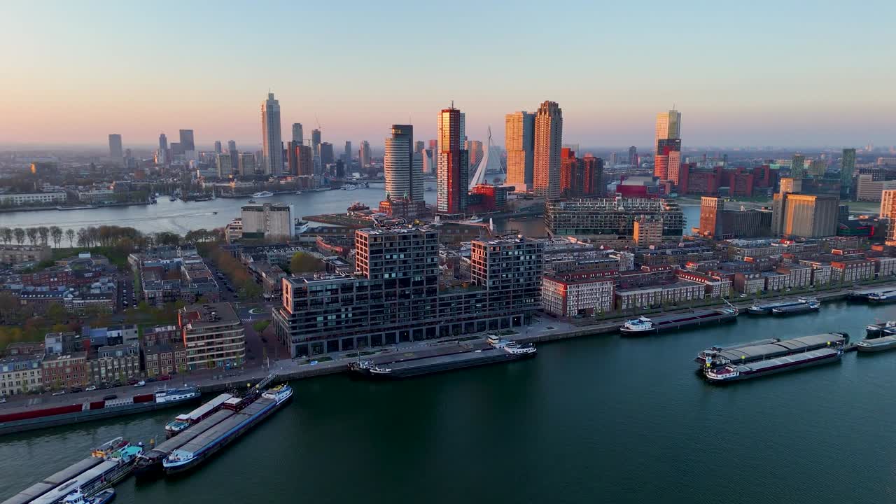 Aerial drone footage of Rotterdam Netherlands showing modern riverfront skyline, Erasmusbrug district, and illuminated buildings reflecting warm sunset light over Maas River and harbor area