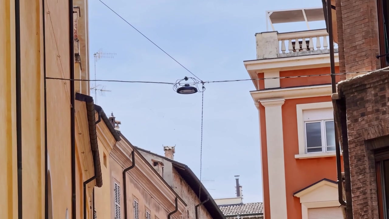 Suspended streetlight and overhead cables span a narrow lane in Parma, Italy, framed by colorful historic buildings and residential facades under a clear blue sky.