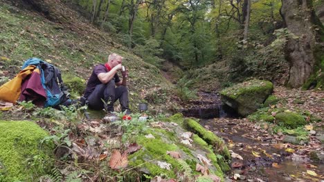 young male hiker enjoys hot tea and snack in forest beside tiny stream