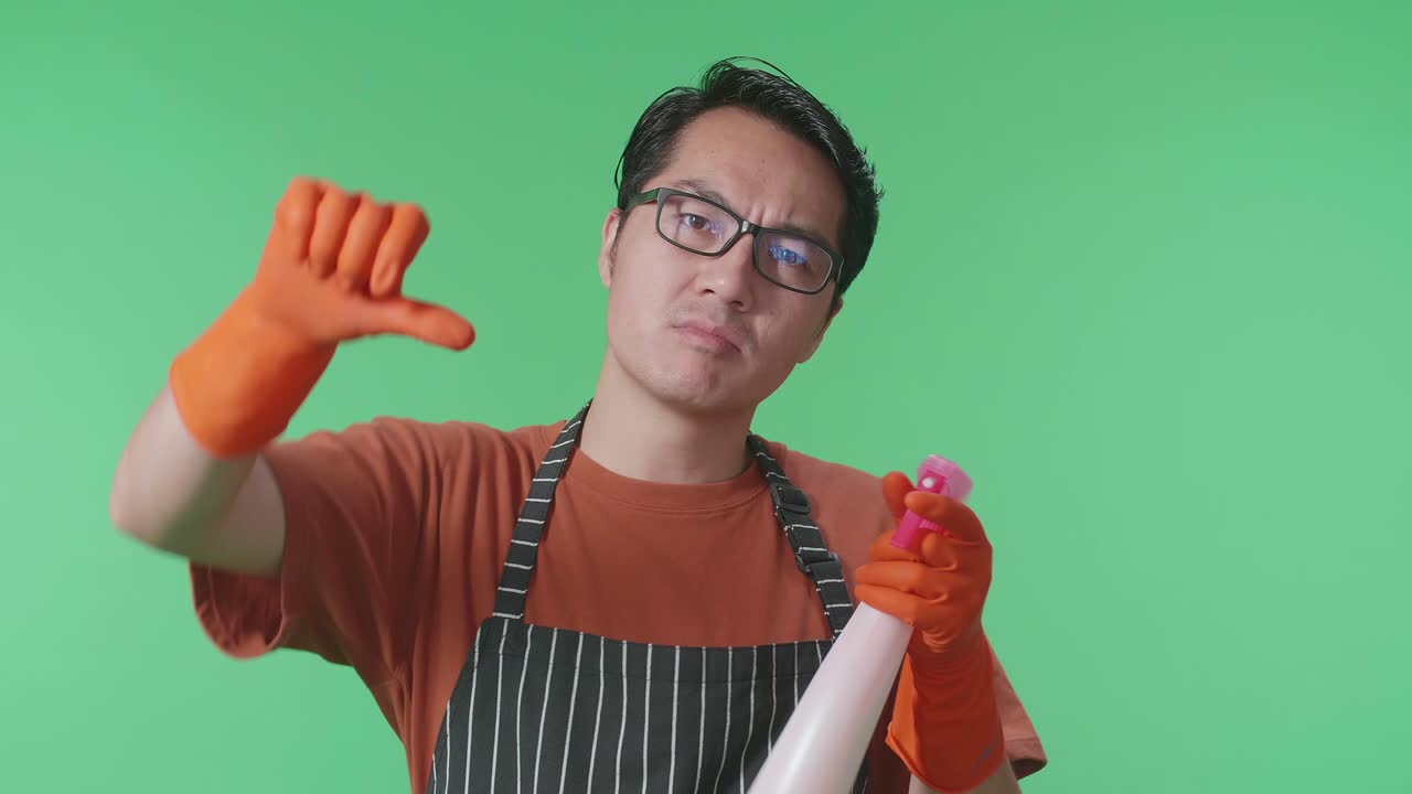 손에 스프레이를 들고 있는 아시안 남성 하녀의 클로즈업 사진. 그 다음에는 손가락을 아래로 내리고 녹색 스크린 스튜디오에서 카메라를 향해 몸짓을 한다.