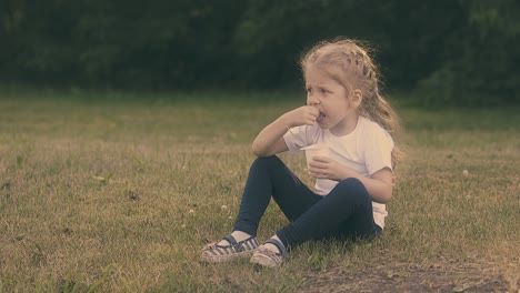 una pequeña dama bonita con cabello rubio come yogur en la hierba del parque