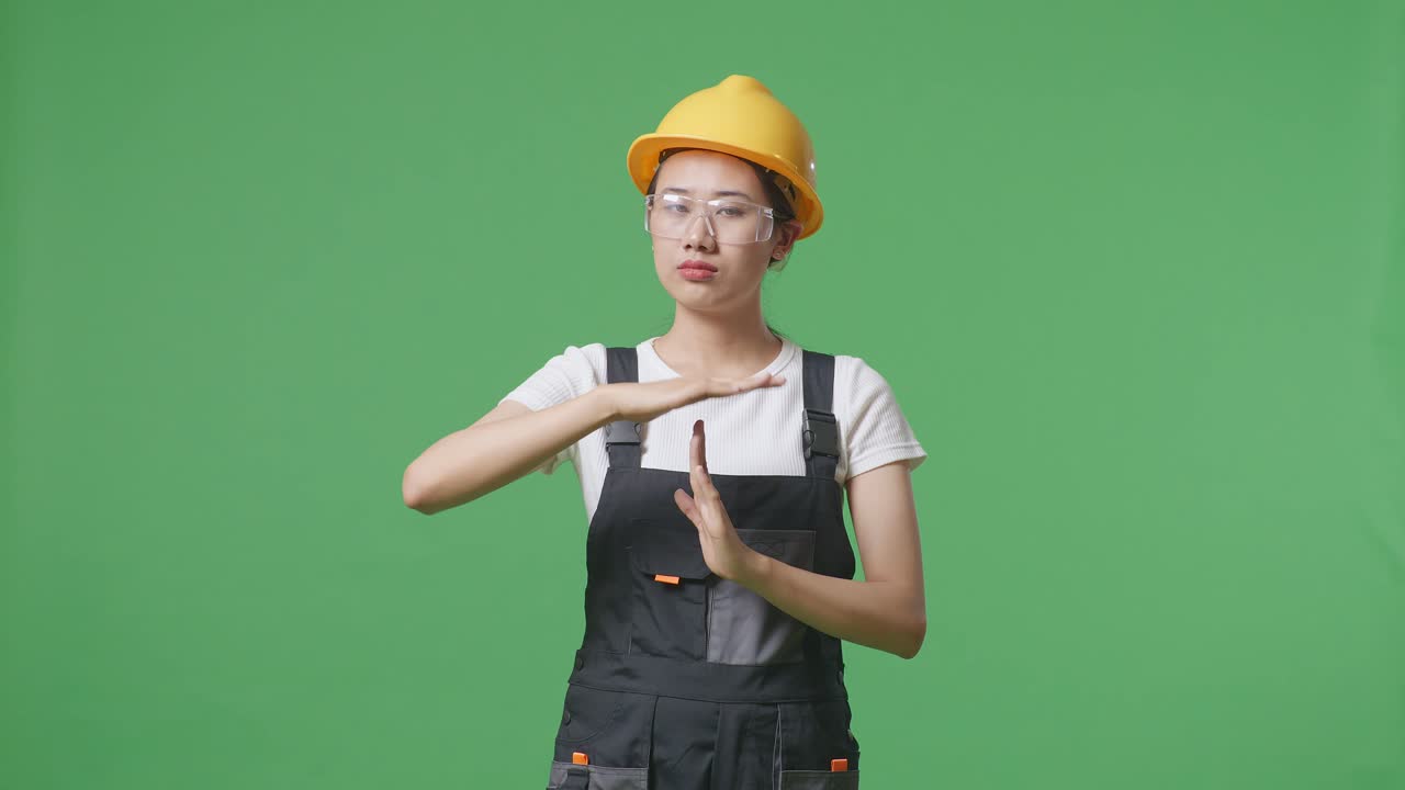 안경과 안전 헬을 착용 한 아시아 여성 노동자가 카메라를 보고 녹색 화면 배경 스튜디오에 서있는 동안 손 제스처를 보여줍니다.