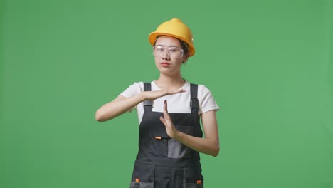 asian woman worker wearing goggles and safety helmet looking at camera and showing time out hands gesture while standing in the green screen background studio