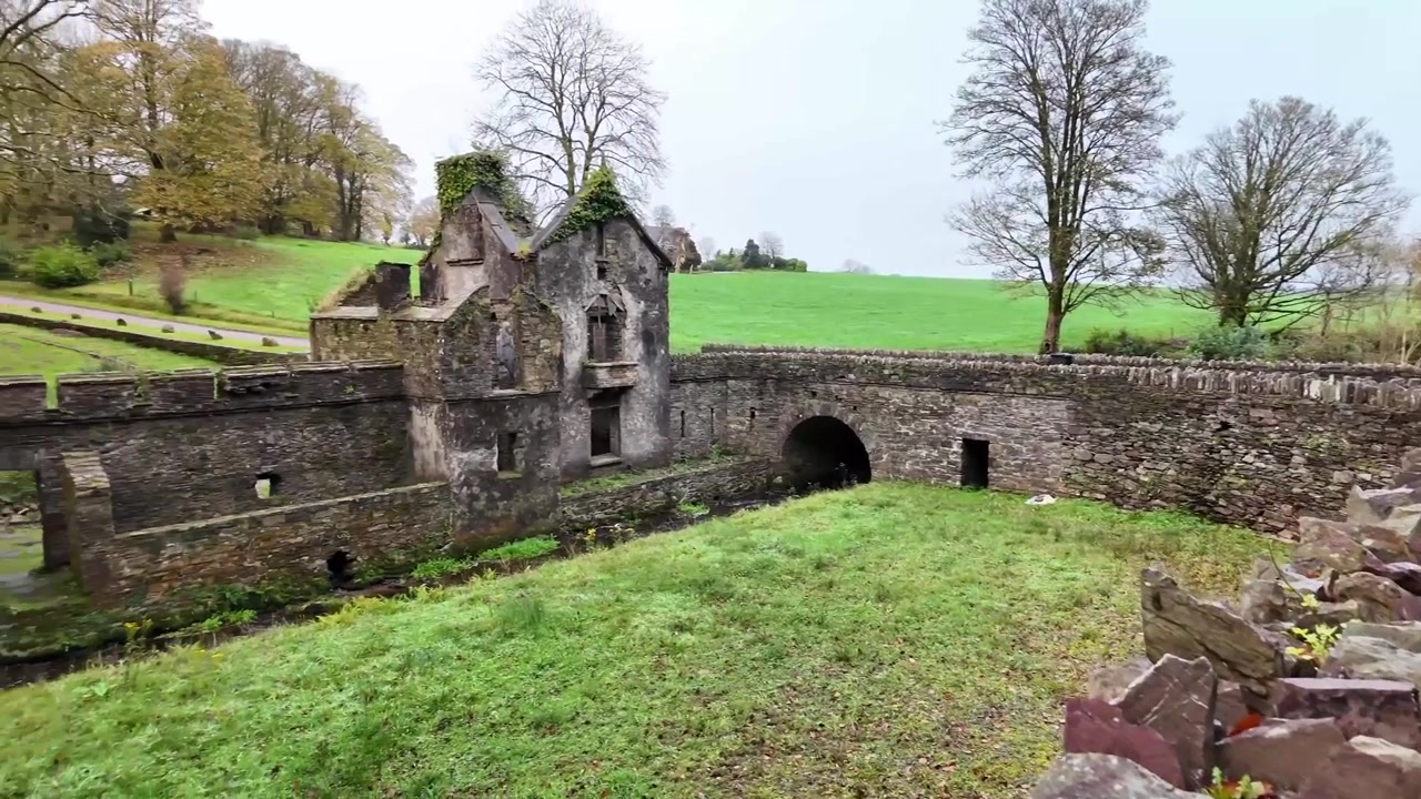 Vej dækket af nedfaldne blade og omkranset af en stenmur. En stenbro over en bæk og ruinerne af et tidligere porthus. Cork County, Irland