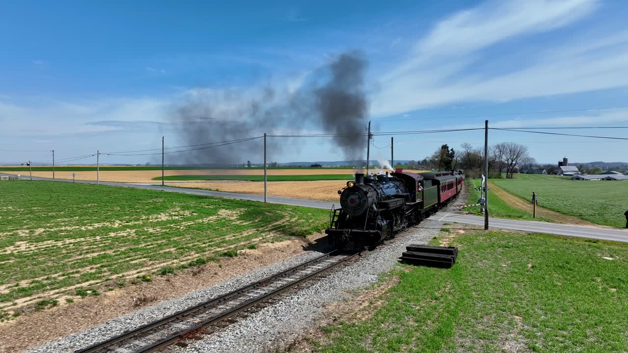 A steam locomotive chugs along the railway, releasing puffs of smoke into the clear blue sky. The train moves through a picturesque rural area with vibrant fields