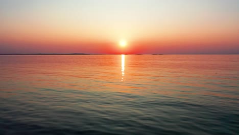 Low-Flying-Drone-View-of-Gorgeous-Ocean-Sunrise-with-Bright-Glowing-Sun-Casting-Colorful-Red,-Orange,-Purple-and-Yellow-Reflections-Over-Peaceful-Rippling-Waves-of-the-Sea-with-Beautiful-Sky-and-Water