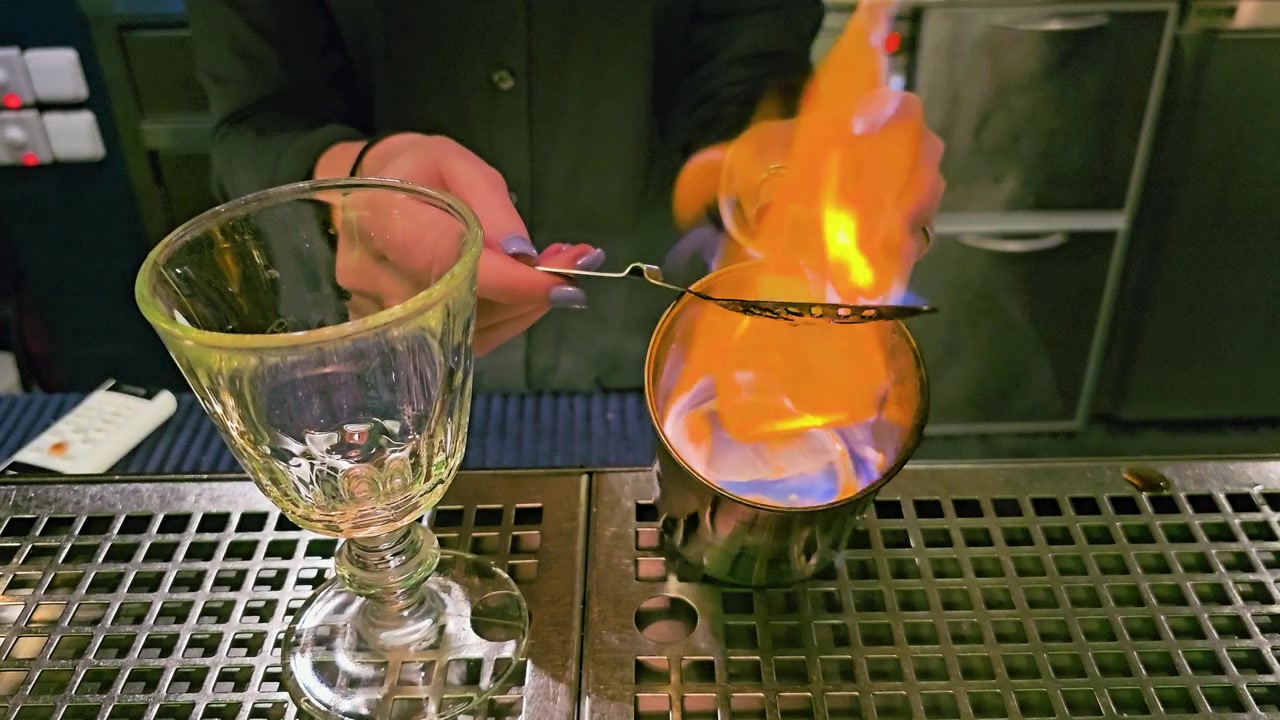 Người pha chế đang cầm một chiếc thìa absinthe có rãnh trên một chiếc cốc kim loại khi ngọn lửa xanh bao quanh một viên đường, cùng với ly thủy tinh cắt, quầy bar inox và ánh sáng lounge xanh mờ ảo.