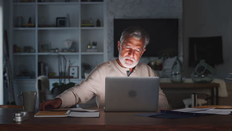 Un-Hombre-Hispano-Mayor-Revisando-Sus-Finanzas-En-Línea-En-Casa-Usando-Una-Computadora-Portátil-Por-La-Noche,-De-Cerca