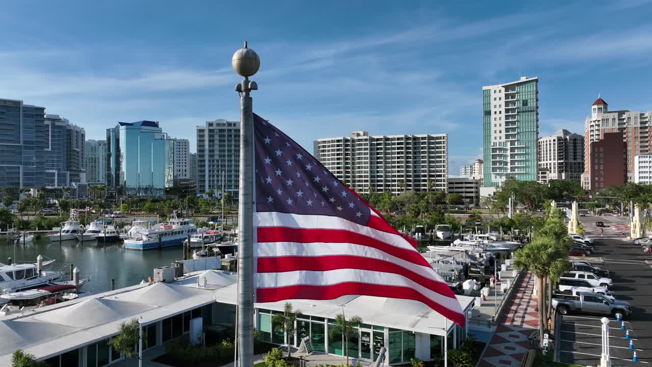 Patriotic America flag on flagpole at marina port of Sarasota city, Florida. Aerial close up orbit shot. Skyline and apartments in background. Luxury yachts and boats in USW country of freedom.