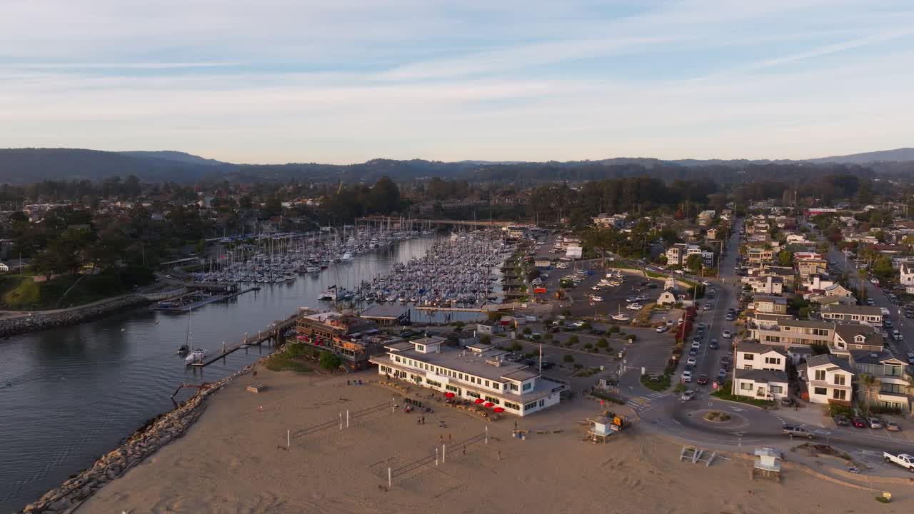 A wide drone shot circling the small craft harbor in Santa Cruz California revealing nearby businesses (Crow's Nest Restaurant and Beach Market) and the Murray St Bridge construction in the background