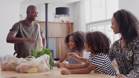 father returns home from shopping trip as mother helps children with homework on kitchen table