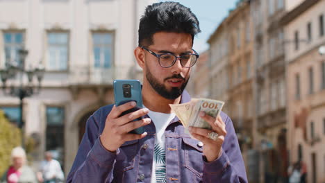 Fröhlicher-Junger-Indischer-Mann,-Der-Geld-Zählt,-Dollar-Bargeld-Verwendet-Smartphone-Rechner-App-In-Der-Stadtstraße