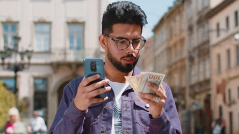 happy young indian man counting money dollar cash use smartphone calculator app in urban city street