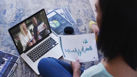 Mixed-race-businesswoman-sitting-on-floor-using-laptop-having-video-call-with-colleagues