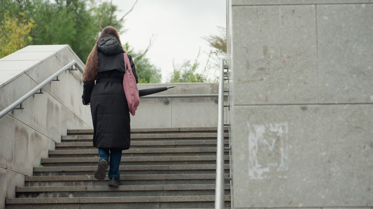 Persona subiendo escaleras de hormigón al aire libre con un abrigo negro y una mochila, llevando una bolsa rosa, ascendiendo una amplia escalera urbana flanqueada por barandillas de hormigón, árboles otoñales nublados al fondo, en soledad