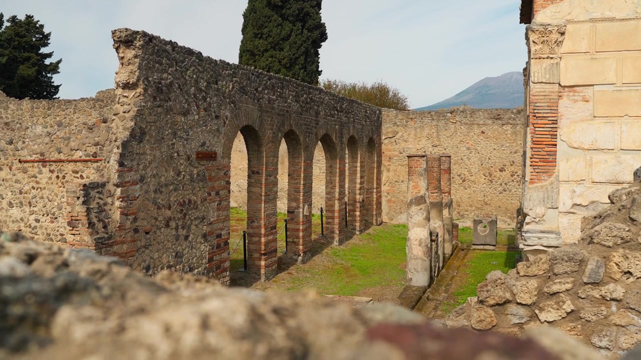Antik Pompei avlu kalıntıları, kemerli taş duvar, tuğla işçiliği, selvi ağacı, korunmuş Roma duvarcılığı ve arka planda Vezüv Yanardağı ile.