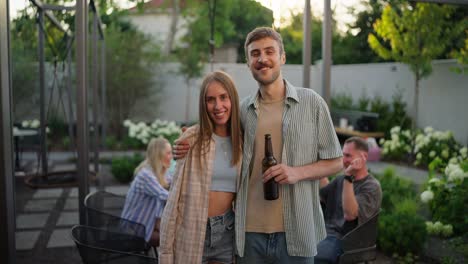 Portrait-of-a-happy-blond-guy-with-his-girlfriend-in-a-blue-top-who-is-hugging-the-guy-and-posing-in-the-courtyard-of-a-country-house